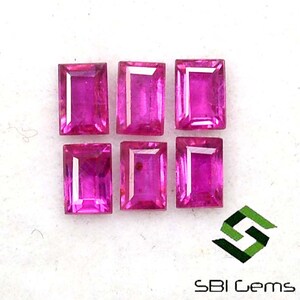 May include: Six pink rectangular cut gemstones, likely rubies, arranged in two rows of three on a white surface.