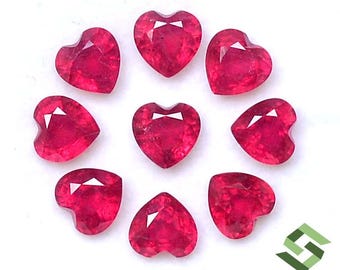 May include: Ten heart-shaped ruby gemstones, each with a deep red colour and a faceted cut. The gemstones are arranged in a circular pattern on a white surface.