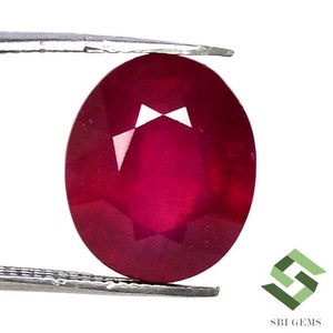 Pode incluir: Uma gema de rubi ovalada de cor vermelha profunda com corte facetado. O rubi é segurado em uma pinça contra um fundo branco.