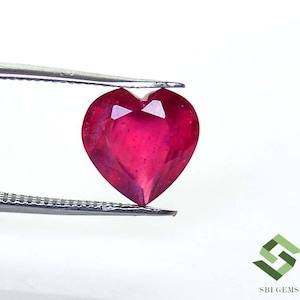 May include: A heart-shaped, faceted ruby gemstone, held by tweezers against a white background. The ruby is a deep, vibrant red color, with visible internal facets. The image is well-lit, highlighting the gemstone's clarity and shape.