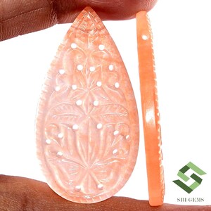 May include: Peach-colored gemstone carved in a teardrop shape, featuring a floral design with small, white dot accents. A side view shows a thin, matching piece with a decorative edge. The gemstone is held by a hand.