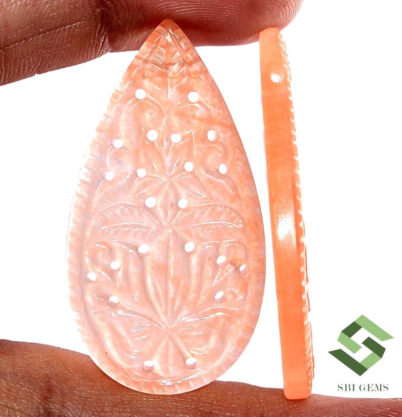 May include: Peach-colored gemstone carved in a teardrop shape, featuring a floral design with small, white dot accents. A side view shows a thin, matching piece with a decorative edge. The gemstone is held by a hand.