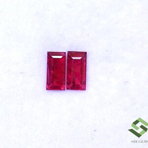 Könnte beinhalten: Zwei rechteckige rote Edelsteine, wahrscheinlich Rubine, werden auf einer weißen Oberfläche gezeigt. Die Edelsteine sind in rechteckiger Form geschliffen mit einer leichten Fase an den Kanten.