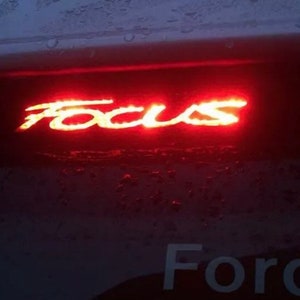 May include: A red illuminated "Focus" emblem on the back of a car with the word "Ford" visible below it.