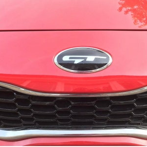 Peut inclure: Une voiture rouge avec une calandre en nid d'abeille noire et une garniture argentée. La voiture a un emblème ovale noir avec les lettres "GT" en blanc.