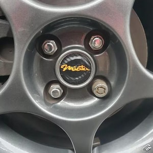 Puede incluir: Primer plano de una llanta de coche gris con una tapa central negra que dice "Miata" en letras doradas. La llanta tiene cinco radios y cuatro tuercas de rueda plateadas.