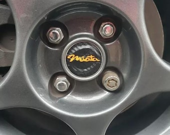 May include: Close-up of a gray car wheel with a black center cap that says "Miata" in gold letters. The wheel has five spokes and four silver lug nuts.