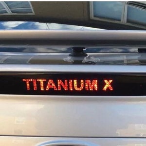 Op de afbeelding: Een zilveren auto met een zwart achterruit en een zwarte achterbumper. De auto heeft een rood "TITANIUM X" embleem op de achterbumper.