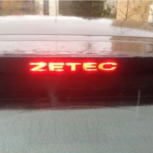 May include: A car's rear window with a red brake light displaying the word "ZETEC" in a bright, yellow-orange hue. The car's body is dark, and the window reflects the surroundings. The image is taken from a close-up angle.