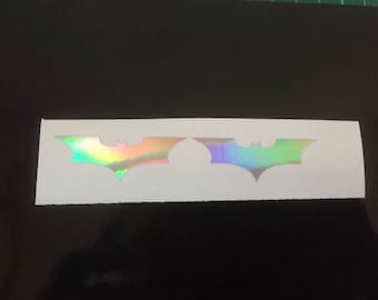 May include: Two iridescent Batman logos on a white background. The logos are a dark silhouette with a rainbow sheen.