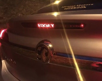 May include: A silver car with a red brake light that says "EDGEY" on the back. The car has a chrome emblem on the back.