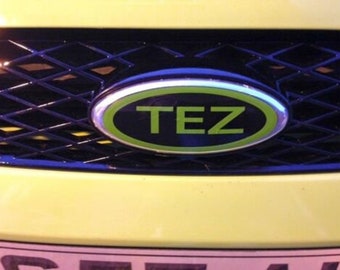 May include: A black and green oval emblem with the word "TEZ" in white letters. The emblem is attached to a black grille with a diamond pattern.