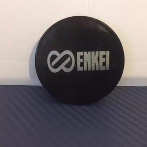 Puede incluir: Un tapacubos negro para una rueda de coche con la palabra "ENKEI" y un símbolo de infinito en plata.