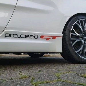 Peut inclure: Une voiture blanche avec un autocollant "pro_cee'd GT" noir et rouge sur le côté. La voiture a des jantes en alliage noir.