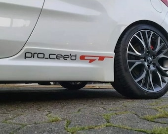 May include: A white car with a black and red "pro_cee'd GT" sticker on the side. The car has black alloy wheels.