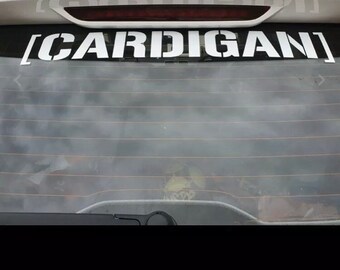May include: A white car with a black rear window. The rear window has a white vinyl sticker with the word "CARDIGAN" in all caps.