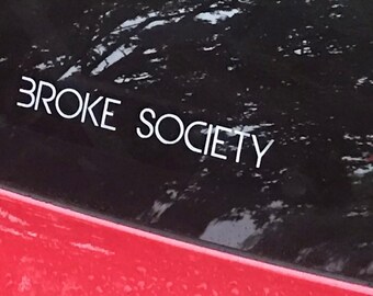 May include: White text on a black background reads "BROKE SOCIETY." The sticker is on a red surface, possibly a car. The image is taken outdoors, with a blurred background of trees.