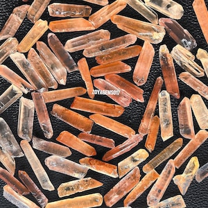 Puede incluir: Una colección de aproximadamente 50 puntas de cristal de cuarzo naranja y amarillo. Las puntas son todas de tamaño y forma similares, y están dispuestas en un círculo suelto sobre una superficie negra.