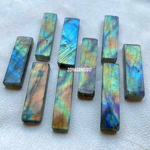 May include: A collection of rectangular labradorite stones. The stones display a range of colors, including blues, greens, and yellows, with iridescent flashes. The stones are arranged on a light-colored surface. The text "zoyagems017" is visible.