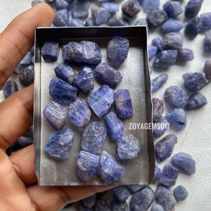 Puede incluir: Primer plano de una pequeña caja de metal llena de piedras preciosas azules-violetas sin pulir. La caja está en una mano, y hay más piedras preciosas sueltas esparcidas alrededor.