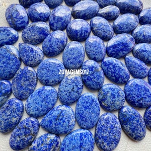 May include: A collection of 25 oval-shaped, polished lapis lazuli cabochons. The stones are a deep blue with white and gold flecks. The cabochons are arranged in a random pattern on a white surface. ZOYAGEMS017 is printed on the surface.
