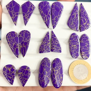 Puede incluir: Una colección de cabujones morados con un patrón de red dorado, dispuestos en pares sobre una bandeja de exhibición blanca. Las formas incluyen triángulos, gotas y óvalos. Una moneda circular se coloca junto a las piedras como referencia.