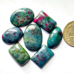 Op de afbeelding: Een verzameling van zes gepolijste cabochon edelstenen met een mix van blauwgroene, groene en magenta-roze insluitingen, gerangschikt naast een Amerikaanse kwartmunt voor schaal. De stenen hebben verschillende ovale en rechthoekige vormen met gladde oppervlakken.