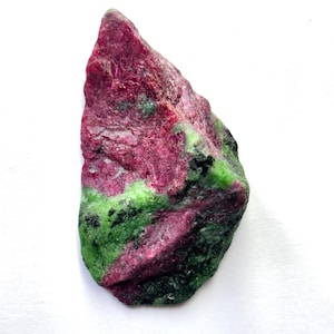 May include: A rough-cut gemstone with a vibrant color palette. The stone features a deep magenta hue, accented by patches of bright green and flecks of black. The natural texture and irregular shape of the stone are prominent.