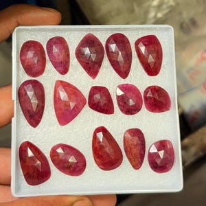 May include: A collection of fourteen faceted, teardrop and oval-shaped red rubies arranged in a white plastic display tray with a foam insert. Each gemstone features a polished surface with geometric facets that reflect light across the deep red.