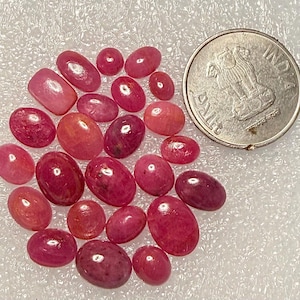May include: A collection of twenty-one oval and round cabochon-cut pink and red gemstones arranged in a cluster next to a circular silver-colored Indian coin for scale. The stones exhibit varying shades of magenta, crimson, and light pink hues.