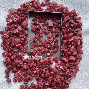 May include: A scattering of raw ruby gemstones, displaying a deep red colour, in various sizes and shapes. The gemstones surround a small, rectangular metal tray. The text "zoyagems017" is visible on the image.