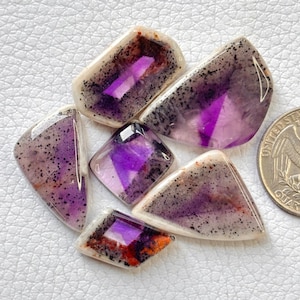 May include: A collection of five polished, faceted gemstones featuring translucent purple and white hues with black speckled inclusions and orange accents, arranged near a United States quarter coin for scale on a white textured surface.