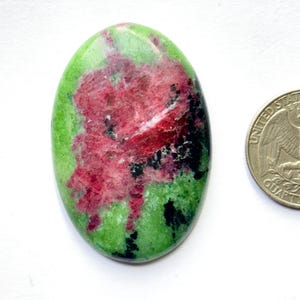 May include: An oval-shaped polished gemstone featuring a vibrant green base with a prominent central patch of deep pink and scattered black inclusions, positioned next to a United States quarter coin for scale reference.