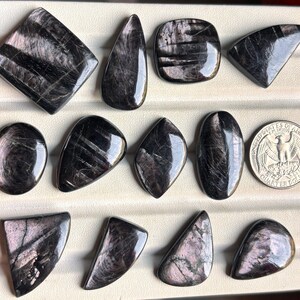 May include: A collection of twelve polished black and dark gray stones with iridescent sheen, arranged in rows. The stones feature various geometric and organic shapes, including squares, ovals, and triangles, displayed next to a US quarter dollar coin.