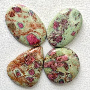 May include: Four polished cabochons featuring a mix of light green zoisite and vibrant magenta ruby inclusions. The stones have smooth, rounded surfaces with natural colour variations, including patches of earthy brown and deep red mineral deposits.