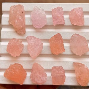 May include: Twelve raw, translucent pink crystals arranged in three rows on a white, ridged display tray. Each crystal features irregular, jagged edges and varying shades of pale to soft rose pink, showcasing a natural, unpolished, and rough texture.