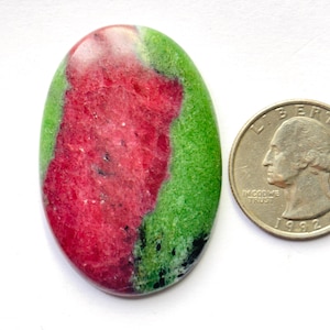 Puede incluir: Una piedra preciosa pulida de forma ovalada que presenta una mezcla vibrante de inclusiones minerales de color rojo magenta intenso y verde brillante con pequeñas motas negras, junto a una moneda de cuarto de dólar estadounidense.