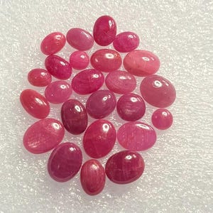 Könnte beinhalten: Eine Ansammlung von einundzwanzig ovalen, polierten rosa und roten Edelsteinen. Die Steine variieren in der Größe und zeigen glatte, abgerundete Oberflächen mit subtilen Lichtreflexionen, angeordnet in einer losen Gruppe auf einer strukturierten weißen Oberfläche.