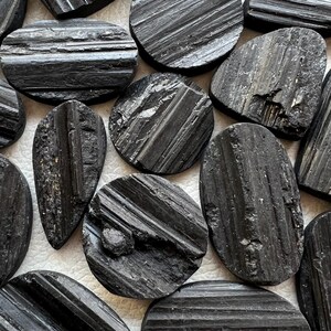 May include: A collection of black tourmaline stones, each with a unique shape and striated texture. The stones are arranged on a white surface.
