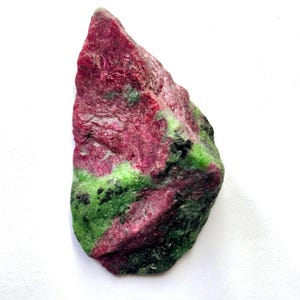 Peut inclure: Un spécimen minéral brut et non poli présentant une combinaison frappante de couleurs rouge magenta profond et vert lime vibrant. La pierre a une surface rugueuse et texturée avec de petites taches d'inclusions noires sombres dispersées.