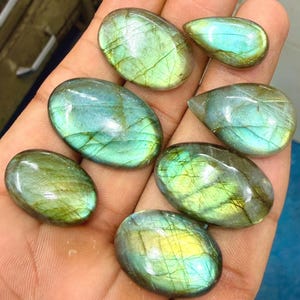 May include: A collection of six polished labradorite gemstones in oval and teardrop shapes. Each stone displays a smooth, reflective surface with iridescent flashes of green, blue, and yellow, featuring natural internal fractures and veining.