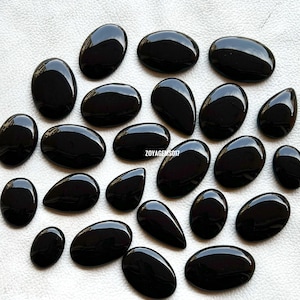 May include: A collection of 20 black gemstone cabochons, some oval shaped and some teardrop shaped, arranged on a white surface.