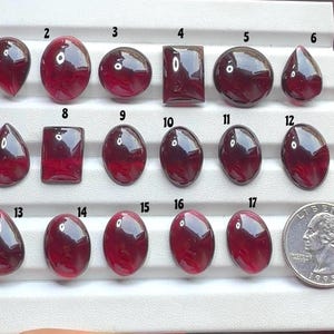 Può includere: Diciassette gemme cabochon lucide, rosso scuro e traslucide, disposte in tre file su un vassoio espositore bianco. Ogni pietra è numerata da 1 a 17. Una moneta da un quarter statunitense è posizionata accanto alla diciassettesima pietra.