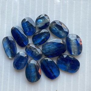 May include: A collection of twelve blue kyanite gemstones, each with a unique, faceted shape. The stones are arranged in a cluster on a white background.