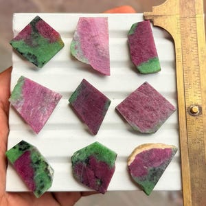 May include: Nine flat, polished slices of ruby zoisite gemstone arranged in a grid. Each piece displays a unique combination of vibrant magenta-pink ruby and deep green zoisite. A brass calliper is positioned on the right, showing measurements in cm.