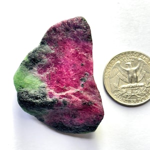 May include: A rough-cut ruby zoisite gemstone with a quarter coin for size comparison. The stone displays a vibrant mix of colors, including deep magenta, black, and green. The coin is silver and shows the image of an eagle.