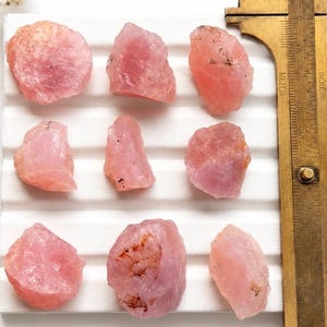 Könnte beinhalten: Neun rohe, rosafarbene Rosenquarzsteine, angeordnet in einem drei-mal-drei Raster auf einer weißen, geriffelten Oberfläche. Ein Messschieber aus Messing liegt an der rechten Seite und zeigt Maße in Zentimetern zur Skalierung der Kristalle.