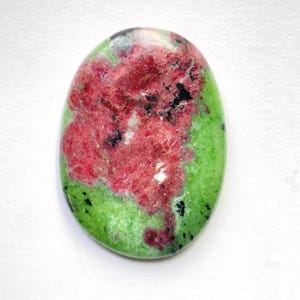 Puede incluir: Una piedra preciosa pulida de forma ovalada que presenta un color base verde vibrante con una mancha central prominente de inclusiones minerales de color rosa intenso y rojo, acentuada por pequeñas motas negras dispersas por la superficie lisa.