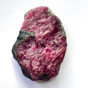 Könnte beinhalten: Ein roher Rubin-Edelstein mit tiefroter Farbe, strukturierter Oberfläche und natürlichen Unvollkommenheiten. Der Stein hat Bereiche in Dunkelgrau und Grün, vor einem weißen Hintergrund.