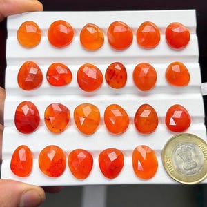 May include: A collection of twenty-three faceted, orange carnelian gemstones arranged in rows on a white display tray. An Indian ten-rupee coin is positioned in the bottom right corner for scale, providing a clear reference for the size of the stones.